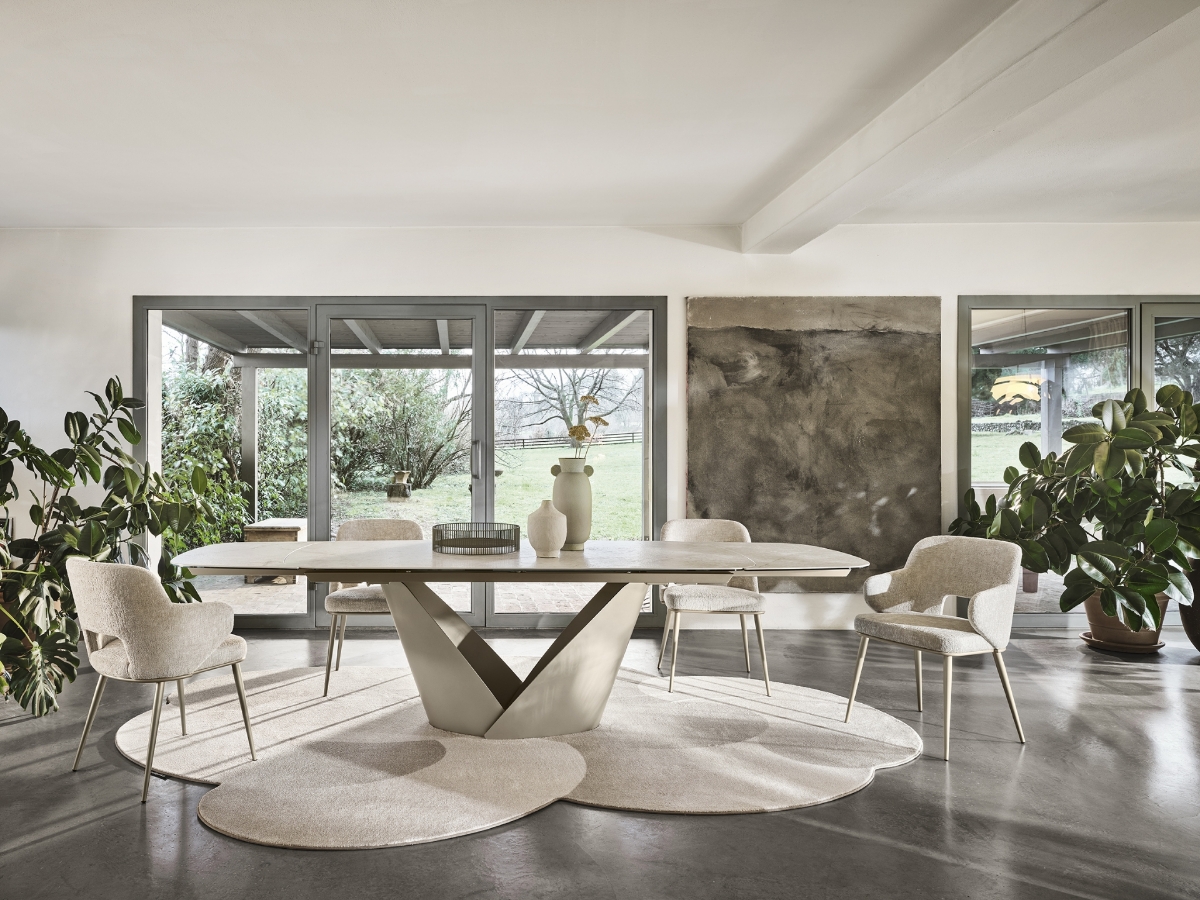 Χαλί σαλονιού Calligaris Curved ακανόνιστο μπεζ κάτω από εκρού κεραμικό τραπέζι με V-βάση και εκρού boucle καρέκλες σε φωτεινό χώρο με μεγάλα παράθυρα και φυτά — Adamopoulos Casa Ideale